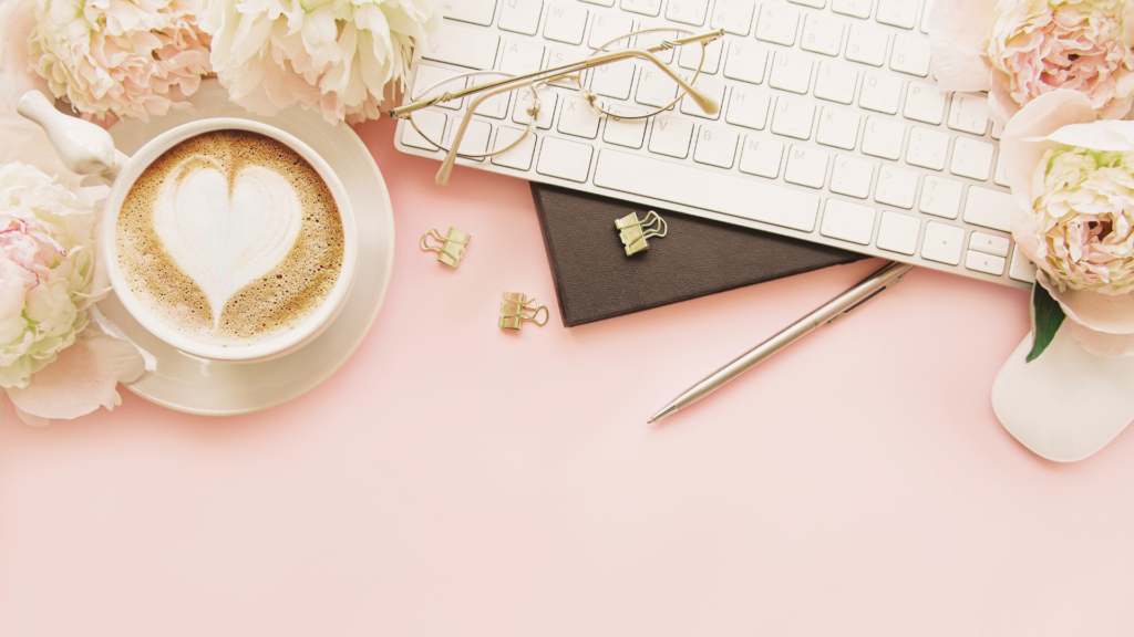 Feminine office space with white keyboard, white and pink peony flowers, white mouse and gold pen, readers and clips on a pink desk